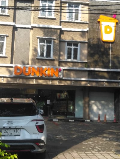 The image depicts the exterior of a multi-story building housing a Dunkin' establishment on its ground floor, situated in the city of Pital, Costa Rica. The building features a large orange "DUNKIN'" sign above the entrance and a prominent, illuminated orange-and-white coffee cup logo with a 'D' on the upper right facade. The upper floors are characterized by light-colored, possibly textured, siding and several rectangular windows with white frames and vertical security bars. In the foreground, a white Hyundai Creta SUV is parked with its rear visible, displaying a license plate "B 2411 KIC" with "10-27" centered below the number. The parking area is paved with interlocking bricks and asphalt, delineated by black and white striped parking barriers. To the right of the Dunkin' entrance, a closed garage-style door is visible, next to three orange traffic cones arranged vertically. Overhead power lines are present across the top of the frame. The scene is observed under daylight conditions, casting shadows on the ground. No individuals are visible in the frame.