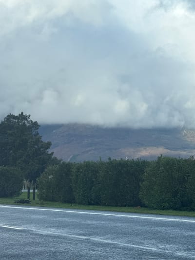 The image captures a scene near Paarl, South Africa. A road runs vertically on the right side of the image, with a white dividing line. Adjacent to the road is a stretch of green vegetation, transitioning into trees. To the left of the trees, a mountainside is visible, partially obscured by low-lying clouds. The sky is predominantly cloudy. The image has been rotated.