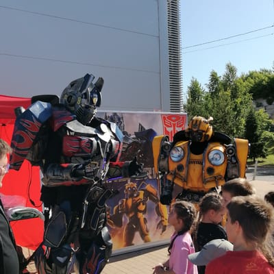 The image captures an outdoor scene in Трубачеевка, Russia, featuring two costumed individuals portraying characters from the Transformers franchise, Optimus Prime and Bumblebee. The individual on the left is dressed as Optimus Prime, wearing a multi-piece costume in dark gray, red, and blue, detailed with red flame patterns and blue illuminated accents. The individual on the right is in a yellow and dark gray Bumblebee costume, designed to resemble vehicle parts, with prominent circular headlight-like elements on the chest.
A group of approximately five to seven children and one adult are observing the costumed characters from the right side of the frame. An adult wearing glasses and a dark jacket is partially visible on the far left, looking towards the Optimus Prime costume. Among the children, one girl in a pink shirt with braided pigtails is looking up towards the Bumblebee costume, while other children are partially visible from behind, including one wearing a red shirt and another with a grey baseball cap.
In the background, a large white banner displays additional Transformers imagery, including an illustration of Bumblebee and the red Autobot faction logo. To the far left, a red fabric canopy is visible. The ground is a paved surface made of light-colored blocks. Behind the scene, a large, light-colored building with distinct horizontal paneling covers a significant portion of the background, with a section of vertical slatted architecture on its right side. Green trees are visible to the right of the building, beneath a clear blue sky. Two thin, dark electrical wires traverse the upper portion of the sky.