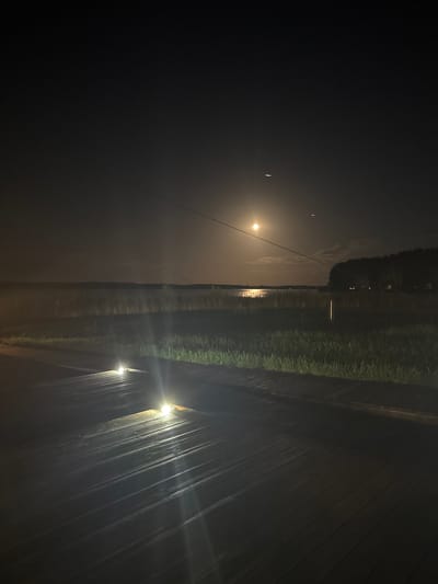 This nighttime photograph depicts a wooden boardwalk illuminated by embedded lights, leading towards a body of water. Beyond the boardwalk, there is a strip of grass followed by a dense line of trees bordering the distant shore. A large, bright moon is visible in the dark sky, reflecting on the water's surface. The overall scene suggests a natural landscape at night, possibly near a lake or river, within the Слабадскі сельскі Савет, Belarus region. There are no people or animals present in the image.