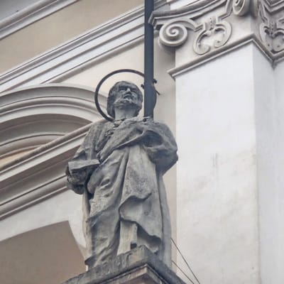 A weathered stone sculpture of a bearded male figure stands against a pale building facade. The figure is depicted wearing a flowing robe and holding an open book in its left hand. A circular halo is visible behind its head. A dark, vertical rod or staff extends upwards behind the figure. The sculpture is positioned on a stone pedestal, part of which is visible at the bottom of the frame. The architectural elements of the building include layered moldings and a decorative scroll motif on a column to the right. The image appears to be taken from a low angle, looking up at the sculpture. This sculpture is located on the exterior of the Church of Santa Maria Assunta in Calcinate, Italy.