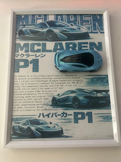 This is a framed poster featuring images of the McLaren P1 hypercar. The poster has a distressed, blue and white aesthetic and displays the "MCLAREN" and "P1" logos prominently in large, blocky blue lettering. There is also Japanese text, "マクラーレン P1," which translates to "McLaren P1." The poster is comprised of three images of the car, with the top image showing the front 3/4 view, a smaller, toy-like model of the car in the middle, and the rear 3/4 view of the car below that. The bottom portion of the poster features a front 3/4 view of the car. The text describes the McLaren P1 as an ultimate hybrid hypercar. The poster is displayed on a wall, and the background is blurred, suggesting an indoor setting. There is no indication of the poster's specific location in Oleśnica, Poland.