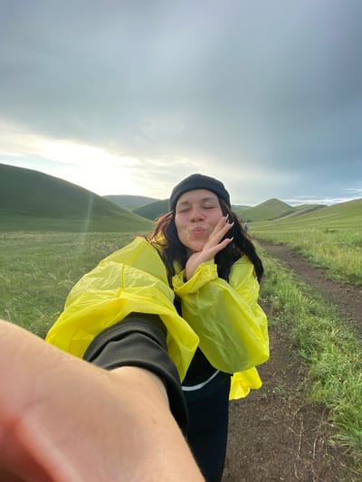A selfie shows a young woman with long, dark hair wearing a black beanie and a bright yellow rain jacket. She is puckering her lips and holding one hand up to her face, with her eyes closed. She is standing on a dirt path in a grassy, rolling landscape. In the background, hills covered in green grass stretch towards a cloudy sky. The overall impression is of someone enjoying the outdoors, possibly on a hike. The location is the city of Donskoe, Russia.