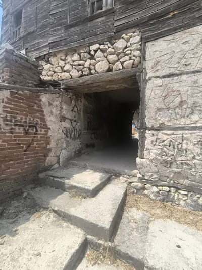 The image depicts an old, weathered stone and wooden structure with an arched opening, likely an underpass or tunnel, leading into darkness. The exterior walls are a mix of rough-hewn stones and aged wooden planks, showing signs of decay and weathering. Graffiti is visible on some of the stone surfaces. In front of the opening, there are a few weathered concrete steps leading up to the entrance. The scene is set outdoors, under what appears to be a clear sky, with dry vegetation scattered around the base of the structure. The architectural style suggests it is an older building, possibly in a historic part of Eğirdir, Turkey.