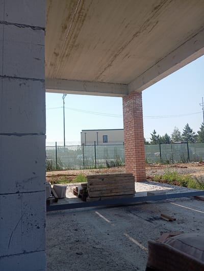 The image shows a construction site in Terema, Russia, on a bright, sunny day. In the foreground, a large concrete column is visible on the left, partially obscuring the view. To the right, a brick column supports a concrete ceiling. Several stacks of wooden planks are placed on the ground, along with some concrete blocks. A metal beam runs across the foreground. In the background, a green metal fence encloses the property. Beyond the fence, there are several modern houses and trees. A tall street lamp with a single light fixture is also visible in the distance. The ground is covered with dirt, gravel, and some patches of grass. The overall scene depicts an unfinished construction project in a semi-rural or suburban area.