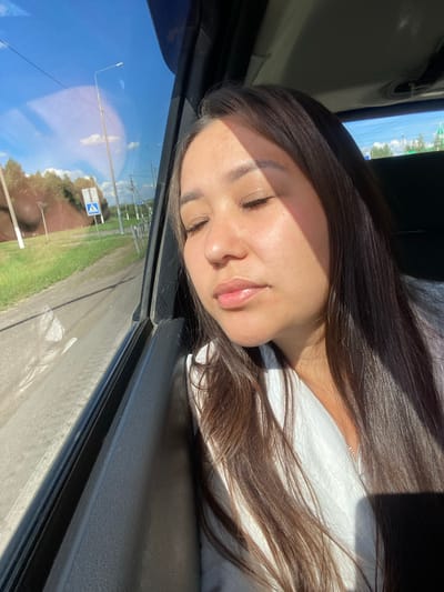 A young woman with long, dark hair is seated inside a vehicle, her eyes closed and her head tilted slightly downwards. Sunlight streams through the window, casting a shadow across her face and highlighting her features. She is wearing a white top. The vehicle's interior is visible, including the door panel and a portion of the window frame. Outside the window, a rural landscape can be seen, with a dirt road, green grass, trees, utility poles, and a blue pedestrian crossing sign under a bright, sunny sky with scattered clouds. The image appears to have been taken in Isyangulovo, Russia, suggesting a journey through a rural area.