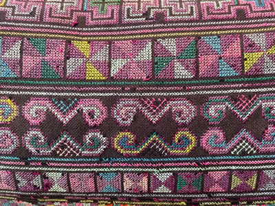 An intricate woven textile displays repeating geometric motifs in horizontal bands. The color palette includes shades of purple, pink, yellow, green, white, and blue. The patterns vary from linear maze-like designs to squares with internal divisions and swirling, wave-like forms. The woven texture and varying stitch densities add depth and dimension. The fabric shows signs of age and wear, with some stitch loss.