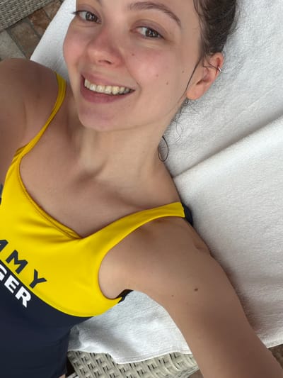 A woman lies on a white towel on a surface that appears to be wicker or similar material. She is wearing a swimsuit that is yellow and navy blue with the word "TOMMY" partially visible. She is smiling and looking directly at the camera. The background is slightly blurred, but suggests an outdoor setting with hints of floor tiles or stone. The location cannot be determined to be Dulepovo, Russia.