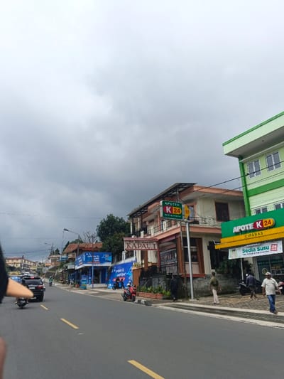 The scene depicts a street in Palayan, Philippines, under an overcast sky. Commercial signage visible, however, identifies "Cipanas." A two-lane asphalt road with a yellow dashed center line extends towards the background.
On the right side of the road, a light green multi-story building features a large sign for "APOTEK K 24 CIPANAS," advertising "Sedia Susu" and "24 JAM" service. Adjacent to it stands a light peach multi-story building displaying another "APOTEK K 24" sign and, below it, a "JUTAH MAKAN KAPAU I" restaurant sign. Further down the street, blue-fronted shops exhibit signs for "Jualan XL" and "Internet Cepat."
Multiple male pedestrians are observed on the right-side sidewalk, including one in a white t-shirt and blue jeans walking away from the camera. Vehicular traffic on the road includes a black SUV and several scooters/motorbikes, some in motion, others parked. A red scooter is specifically parked on the shoulder near the peach-colored building. Utility poles and overhead power lines are visible along the street. A blurred, dark object partially obscures the immediate foreground on the left.