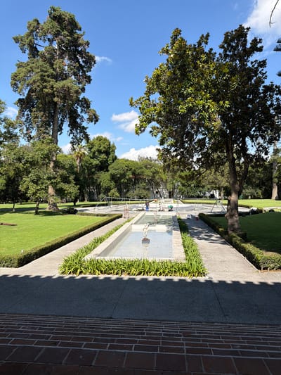 This image captures a vibrant outdoor scene within a well-maintained garden or park, likely in San Lucas el Grande, Mexico. The perspective, rotated 90 degrees clockwise, looks down a long, rectangular water feature or decorative pool that stretches into the distance.
The water feature, made of a light-colored material, contains several striking white, skeletal-like sculptures or abstract figures, resembling human forms, positioned at various points along its length. A metallic railing runs alongside one edge of the feature. To its right, a concrete pathway is bordered by manicured green shrubs and planters. This pathway is partially covered by a sharp, jagged shadow, indicating a strong light source and suggesting late morning or early afternoon. Further to the right, a textured brick wall or pathway extends along the entire right side of the frame.
To the left of the water feature and path are lush green lawns and a variety of mature trees with dense foliage, contributing to the park-like atmosphere. Two prominent trees stand in the foreground and mid-ground on the left, with their full canopies visible against the clear blue sky, which features scattered white clouds. Several people are seen near the water feature, observing the sculptures or strolling along the paths. One individual in a green shirt is leaning over the edge of the pool, while another in a blue shirt stands further back. The scene conveys a peaceful and pleasant outdoor environment. No discernible text is visible in the image.