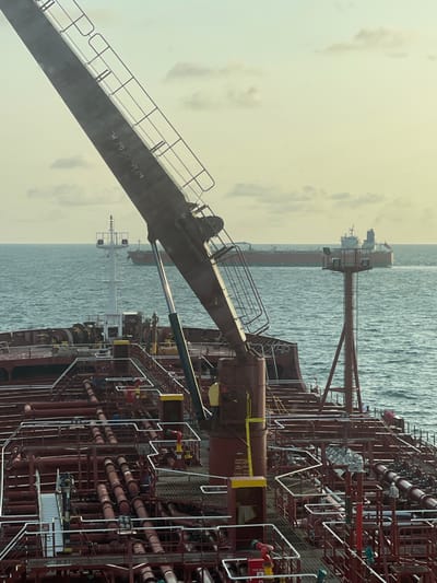 The image displays an offshore scene near Orimedu, Nigeria, viewed from the deck of a large vessel, likely an oil tanker. In the immediate foreground, a substantial portion of the vessel's deck is visible, characterized by an intricate network of dark red pipes, valves, and white metal railings. A prominent dark-colored crane arm, marked "SW 10 TON," extends diagonally upwards from the center-left, supported by a hydraulic cylinder. Various deck structures, including a white mast with lights and a red, multi-tiered navigational tower, are also present on the foreground vessel. In the mid-ground, the ocean surface exhibits moderate choppiness under an overcast sky with scattered clouds. On the distant horizon, another large, red-hulled vessel, resembling an oil tanker with a white superstructure, is visible, appearing to be underway. No individuals are discernible on the visible decks.