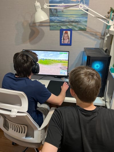 Two males are situated in an interior setting. One is seated at a desk wearing headphones and playing a video game displayed on a computer monitor. A second male stands behind him, looking at the screen. A computer tower with illuminated blue components is on the desk. There are two lamps, one adjustable and one fixed, and wall art that includes a framed photograph of a child. The walls are wallpapered with a subtle textured pattern. The location cannot be confirmed as Dzerzhinsky, Russia, based solely on this image.