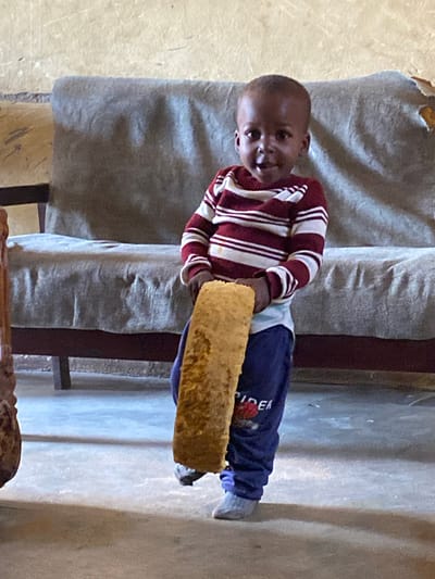 A young child, likely a boy given the clothing and short hair, stands in an indoor setting holding a large, elongated piece of fried dough or bread. The child wears a striped long-sleeved shirt in red and white and dark blue pants with a red graphic and white text on the left leg. Light grey socks cover the child's feet, and one foot appears to be wearing a dark shoe. The background features a neutral-colored couch with a textured surface and a light-colored, slightly worn wall. The floor is a light grey, concrete-like material. The lighting suggests an indoor environment, possibly in a home or communal space. The image captures a moment of the child holding the food item, looking towards the camera with a slight smile. The scene appears to be located in Panyam, Nigeria, based on the provided context.
