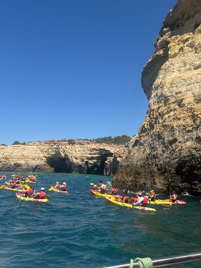 A group of kayakers paddle in formation near a towering cliff face under a clear blue sky. The kayakers are predominantly wearing red life vests and are spread across the choppy, blue water. The cliff, a prominent feature of the landscape, shows layers of weathered rock and features several inlets or caves. The scene appears to be set in the coastal region of Lagoa e Carvoeiro, Portugal, known for its dramatic rock formations and sea caves.
