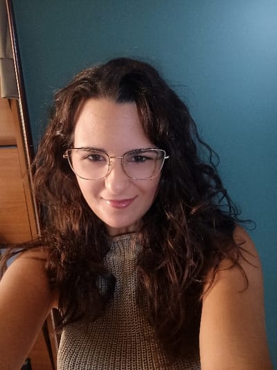 A portrait-oriented image depicts a young adult female positioned centrally, looking directly at the camera, from an indoor setting within San Marzano sul Sarno, Italy. The subject has long, dark brown, wavy hair that frames her face and extends over her shoulders. She wears gold-toned, cat-eye style eyeglasses with a subtle pattern on the upper rims. Her facial expression is a faint, closed-lip smile, and her lips appear glossy with a light pink tint. She is dressed in a sleeveless, light grey or beige knitted top featuring a high, mock-turtleneck collar. The background consists predominantly of a plain teal-colored wall. To the far left, a portion of a wooden furniture item, appearing to be a cabinet or wardrobe, is visible. The lighting is even and illuminates the subject from the front.