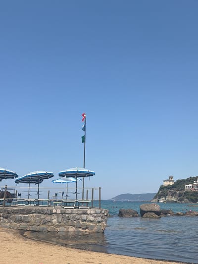 This is an outdoor, horizontal, medium shot of a beach in Castiglioncello, Italy. In the foreground, there is a sandy beach with some debris. A stone wall borders a raised platform with several blue and white striped umbrellas and lounge chairs. The sea is visible behind the platform, with rocks and a few swimmers. In the background, a hill is covered with lush green trees and houses, one of which is a prominent building with a tower. The sky is clear and blue. A flagpole with three colored flags is also visible on the platform.