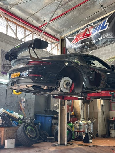 An interior view of an automotive workshop in Azor, Israel, depicts a black Porsche 911 GT3 RS elevated on a red hydraulic lift. The vehicle, bearing Israeli license plate 592-27-501, has its wheels removed, exposing the brake rotors and red calipers; the rear right brake assembly is prominently visible. The car features a large, dark carbon fiber rear wing with distinctive endplates and "Arbel Racing" decals on its side window.
The workshop's ceiling is covered with metallic foil insulation, traversed by red pipes, and an "AMSOIL - The First in Synthetics" banner is suspended. The walls consist of concrete blocks, and the floor is tiled. Scattered around the lift's base are various automotive items, including a pair of wheels, one with a green center lock, a white Porsche-branded shopping bag, blue plastic bins, an oil drain system, and assorted tools and parts. Natural light enters from an upper window on the left, supplementing artificial lighting. No individuals are present in the scene.
