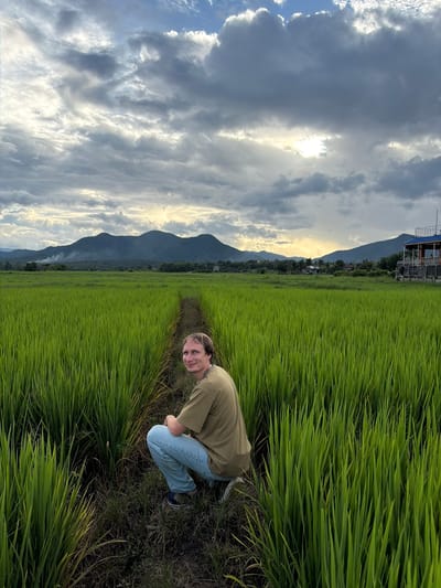 A man is crouching in a rice paddy in Ban Kio Lae Pa Pao, Thailand. The rice paddy is lush green and stretches into the distance, with a path cutting through it. In the background, there are mountains and a building with a blue roof. The sky is cloudy with patches of sunlight breaking through. The man is wearing a green t-shirt and light blue jeans. He is looking at the camera and smiling.