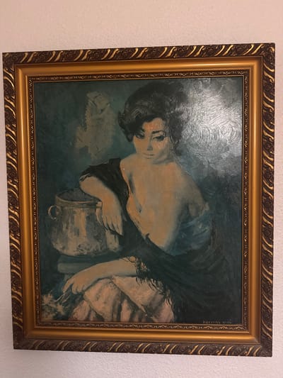 A portrait painting in a golden frame depicts a woman with dark, voluminous hair and a pensive expression. She is seated, draped in a dark shawl that reveals her shoulders and chest. Her right arm rests on a large, metallic pot. In her left hand, she holds a few slender, light-colored objects, possibly feathers or reeds. The background is a dark, textured teal. A signature, "TREINAS VIVO," is visible in the lower right corner of the painting. The painting is displayed on a white textured wall, suggestive of an indoor setting. The image does not provide any specific details or context that would allow for the identification of its location within the city of Santa Pola, Spain.
