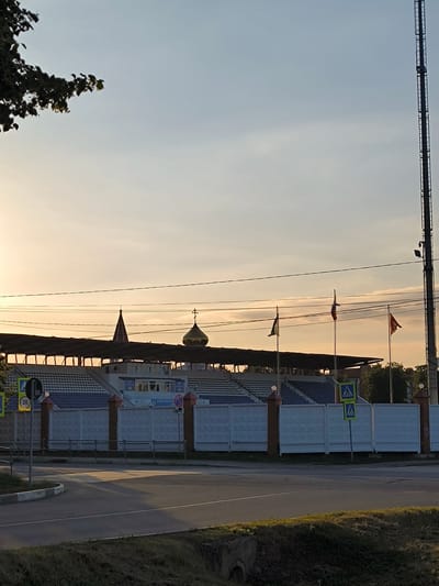 This is a medium-distance, horizontal shot of a stadium in the city of Serebryanye Prudy, Russia, taken at dusk. The stadium features tiered seating under a covered roof. In front of the seating, there is a white fence with decorative pillars. Behind the fence, three flags are visible, one of which appears to be the Russian flag. To the right of the stadium, a tall tower with lights is present. In the foreground, there is a road with a pedestrian crossing and traffic signs, including a speed limit sign showing "20". The sky is a soft orange and yellow due to the setting sun, with some clouds present. A tree branch with leaves is visible in the upper left corner of the frame. The overall scene suggests an outdoor sports or event venue during the evening.