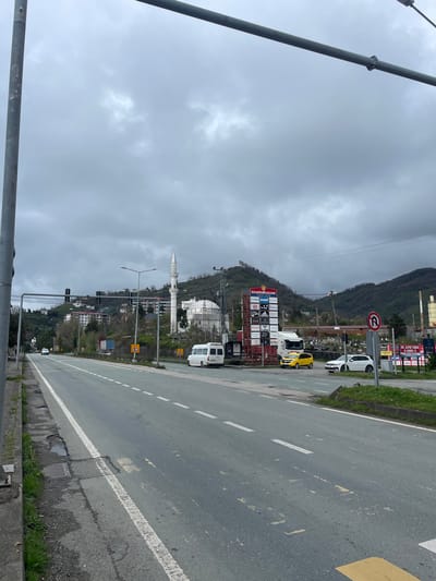 The image shows a street scene in Kemalpaşa, Turkey. On the right, a gray asphalt road is visible with white lane markings and a yellow curb. Parked along the side of the road are several vehicles, including a white van, a yellow car, and another white car. Beyond the parked vehicles is a commercial area with large signs for various businesses, including Istanbul bazaar, Mavi, Adidas, and Flo. Behind the commercial area, a hillside with trees and vegetation rises. On the hillside, a white mosque with a prominent minaret is visible. The sky is overcast with heavy gray clouds. A metal railing is visible in the foreground on the left. Overhead power lines run across the scene.