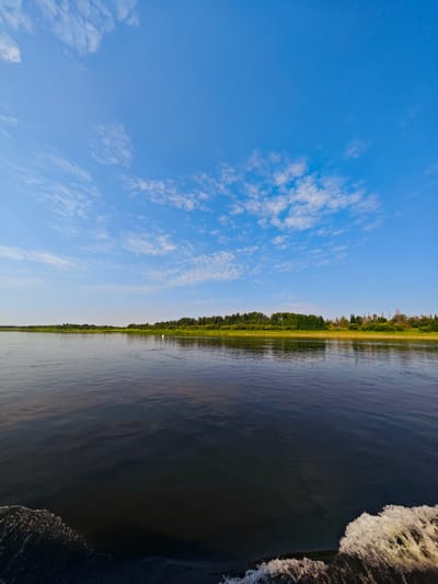 The image depicts a wide, dark river on a clear day. A dense line of green trees and vegetation forms the distant shore. The sky is a vibrant blue, with scattered white clouds. In the middle distance on the water, there is a single white object that appears to be a bird or a buoy. The foreground shows the wake of a boat, with foamy white water churned up. The scene is serene and natural, suggesting a peaceful setting. This could be the Oka River in the vicinity of Novonazimovo, Russia, known for its scenic beauty and recreational opportunities.