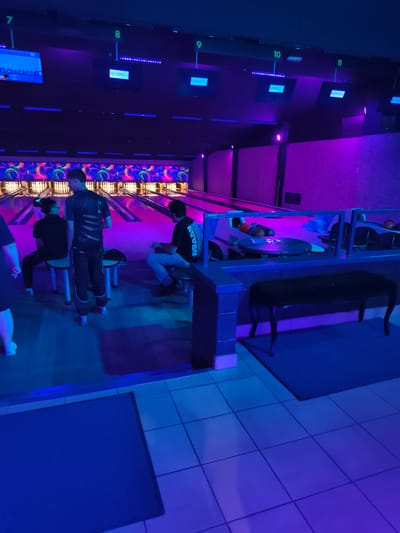 The image depicts an indoor bowling alley with multiple lanes illuminated by purple and blue neon lights. Several individuals are present in the scene, with some seated and others standing. One person is wearing a dark t-shirt with white lettering that appears to be a brand name. Another person is wearing a t-shirt with a graphic design. The bowling lanes are visible in the background, with bowling pins set up. Overhead, screens display scores and the "QUBIC" logo. The flooring consists of light-colored tiles. The environment is characteristic of a modern entertainment venue. The location is identified as Chelles, France.
