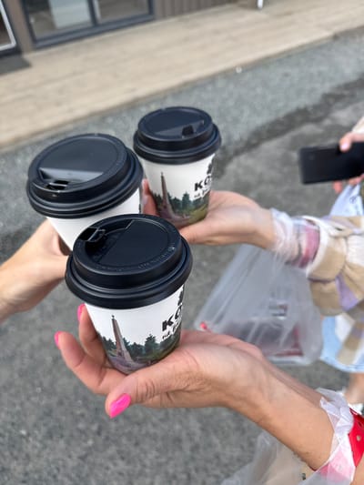 Three to-go coffee cups with black lids are held by three individuals, forming a toast. The cups have white paper sleeves featuring a picture of a monument and trees, along with the text "KO". One individual's hand, with bright pink painted nails, is holding a cup in the foreground. To the right, another person's hand is visible holding a smartphone. In the background, a plastic bag with items inside and a person wearing a plaid shirt are partially visible. The scene appears to be outdoors on asphalt, with a building and its entryway visible in the far background. The specific location in the city of T/B Seminskii Pereval, Russia is not identifiable from the visual information present in the image.