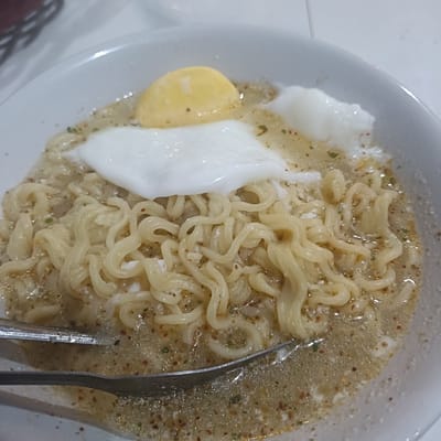 A white bowl filled with noodles in broth and two eggs, one with a hardened yolk and one soft-boiled, rests on a white surface. A set of metal tongs is visible in the lower left portion of the bowl. The location is likely Cangkring, Indonesia.