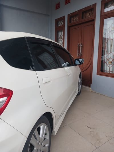 A white hatchback car is parked in front of a house with double wooden doors and two windows. The car is angled with its rear towards the viewer, revealing the passenger side. The house has a light gray wall and a tiled floor. The scene appears to be in Geudonggeudong, Indonesia.