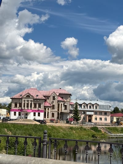 This image depicts a view of buildings in Pereslavl-Zalessky, Russia, situated along a waterfront. In the foreground, a dark metal fence with intricate scrollwork runs horizontally across the lower portion of the frame, partially obscuring a reflective body of water. The water's surface shows ripples and reflections of the surrounding environment.
Behind the fence, a grassy embankment slopes upwards towards a row of buildings. The buildings are of varying architectural styles. The building on the left is a multi-story structure with a light-colored brick facade and a prominent pink tiled roof. It features numerous windows and some visible air conditioning units. A pink stripe with text, likely signage, runs along the lower portion of this building. Adjacent to it is another building, also multi-story, with a beige facade and a silver-colored pitched roof with multiple dormer windows. This building has a more modern appearance. Further to the right, another building with a red roof is visible, along with part of a yellow construction vehicle.
The sky above is a bright blue, dotted with large, fluffy white cumulus clouds, creating a dynamic and somewhat dramatic contrast with the scene below. A few distant trees are visible beyond the buildings, suggesting a semi-urban or suburban setting. The overall impression is that of a sunny day with dramatic cloud formations over a town by the water.