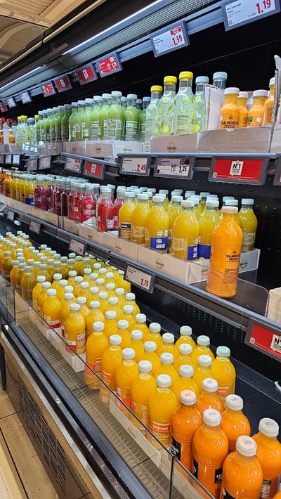 The image shows a refrigerated display case in a supermarket in Cuarte de Huerva, Spain, filled with various bottled juices. The display features multiple shelves stocked with rows of clear and colored glass bottles, predominantly containing yellow, orange, and green liquids. Some bottles have visible labels indicating flavors such as "Piña Ananas" (Pineapple) and "Naranaja/Fresa" (Orange/Strawberry). Price tags are displayed on the front edge of the shelves. The lighting in the supermarket is bright, and the overall environment appears clean and organized. There are no people visible in the image.
