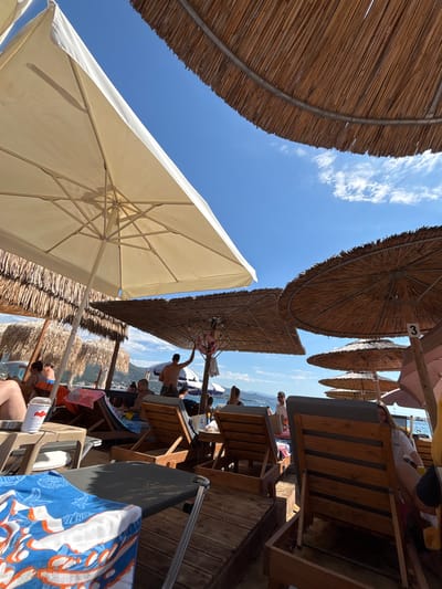 The image depicts a sunny day on a beach in Νέα Ηρακλείτσα, Greece, viewed from beneath a large white beach umbrella and several thatched umbrellas. In the foreground, a blue and white patterned beach towel lies on a wooden deck, with a small table to its left holding a coffee cup. Behind this, rows of empty and occupied wooden lounge chairs are visible, many with patrons relaxing or walking. Several people are scattered throughout the scene, some seated in chairs, others standing. In the background, the blue sea meets a distant, hazy coastline with mountains under a clear blue sky dotted with wispy clouds. The overall atmosphere is that of a leisurely beach resort.