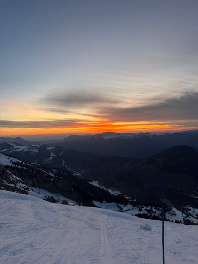 The image shows a mountain landscape at sunset, likely taken from a high vantage point. A snowy ski slope occupies the lower right portion of the frame.  In the distance, dark mountain ranges are visible. The sky is ablaze with colors ranging from pale grey to fiery orange and gold. A blue rope stretches across the scene from right to left. The location is possibly in La Giettaz, France.