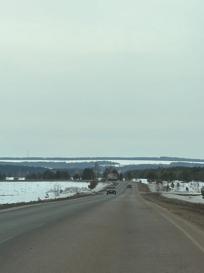 The scene depicts a two-lane asphalt highway extending into the distance within the vicinity of Povarenki, Russia. The sky is uniformly overcast. The road surface is dark, appearing damp or worn, with a single dashed white line marking the center. Snow-covered ground with patches of exposed earth and sparse vegetation, including small coniferous and bare deciduous trees, borders both sides of the highway. A dense line of dark coniferous trees forms a midground horizon. Beyond this, a wide, flat expanse of white, consistent with snow-covered land or a frozen body of water, stretches across the background. A faint, tall, slender, dark tower structure is visible on the left side, rising from this white expanse. A distant, dark tree line marks the far horizon. Four vehicles are observed traveling away from the viewer: a dark sedan in the foreground, a lighter-colored sedan further ahead in the same lane, a white semi-truck with a red cab and red lettering on its trailer positioned in the left mid-distance, and another lighter-colored car on the right side of the road in the mid-distance.