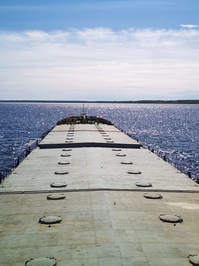 The image shows a view from the deck of a vessel, possibly a barge, on a body of water. The deck is light gray, flat, and features rows of circular access hatches. A small structure is visible at the front of the deck, with a thin, vertical antenna extending upwards. The water is dark blue and choppy, with reflections of sunlight causing it to shimmer. A treeline is visible on the horizon across the water. The sky is pale blue with scattered white clouds. The scene is most likely in a city of Koprino, Russia.