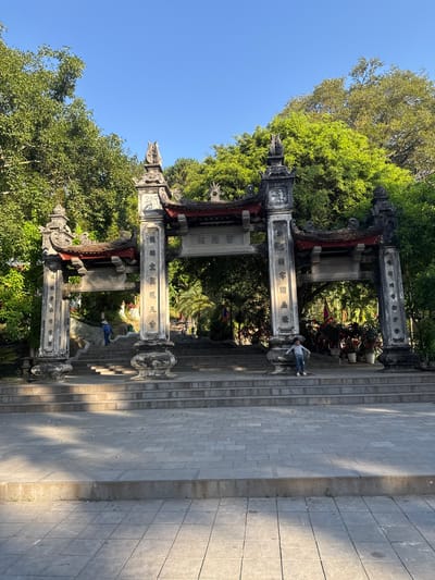 The image shows an ornate, stone entrance gate situated in Lao Cai Ward, Vietnam, with a steep staircase leading up to it. The gate consists of multiple pillars adorned with carvings and text, framed by lush greenery and blue sky. Two people stand within and behind the gate, one near the top of the staircase and the other further down. The staircase is made of rectangular stone blocks, with the paved ground extending to the right. The scene is bathed in sunlight.