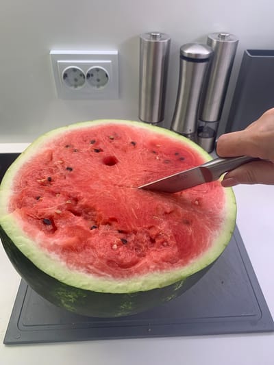 A half watermelon is being cut by a knife held by a person's hand in what appears to be a kitchen. The watermelon is red with black seeds and a green rind, resting on a grey cutting board. In the background, stainless steel salt and pepper grinders are visible, along with a white electrical outlet. The overall scene is a close-up, indoor shot.