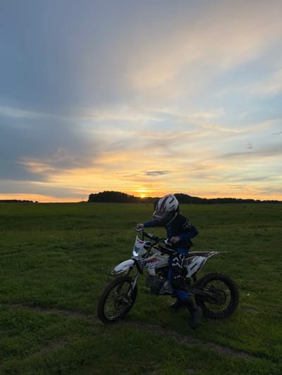A person wearing a helmet and blue riding gear is seated on a white dirt bike in a grassy field. The dirt bike is angled towards the right side of the image. The background features a sunset with orange and yellow hues blending with the sky. A line of trees is visible along the horizon. The photo appears to be taken in a rural setting.

Based on visual elements, the location in the city of Новочувашский, Russia cannot be determined.