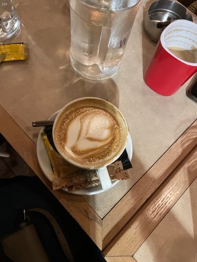 This top-down image captures a cafe table in Chaidari, Greece, adorned with several items. The central focus is a white ceramic cup of latte, featuring a distinct heart-shaped latte art in its foam. The cup rests on a matching saucer, which also holds several brown paper packets labeled "Cane Sugar" from "Lupus Natura" and a metal spoon.
To the left of the coffee, a large, clear glass, covered in condensation and likely containing water, is visible. Near the glass, a small yellow packet of sugar is placed. In the upper left portion of the image, a bright red plastic cup lies on its side next to a metal ashtray containing ash and cigarette butts. A dark mobile phone is partially visible at the top center. In the bottom right corner, part of a light brown or beige handbag is seen against a dark background, suggesting the presence of an unseen individual.
The table surface is a light, textured material, possibly faux leather, framed by a wooden border with a herringbone or similar geometric pattern. The lighting is warm and ambient, indicative of an indoor setting. The arrangement of items suggests a casual break or social gathering.