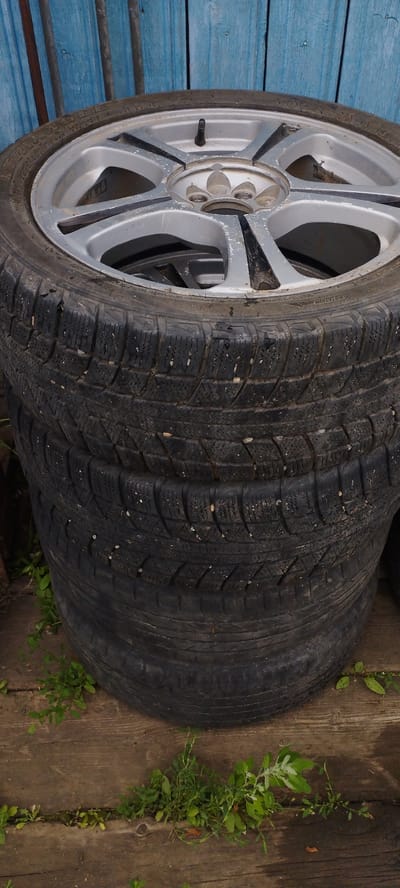A stack of four used car tires is shown in front of a weathered blue wooden fence, with the top tire having a silver rim. The tires are stacked vertically on a wooden surface that is partially covered by small green plants. The scene appears to be outdoors, possibly in a yard or storage area in the village of Pokosnoye, Russia. No people are visible in the image. The lighting suggests it is daytime.