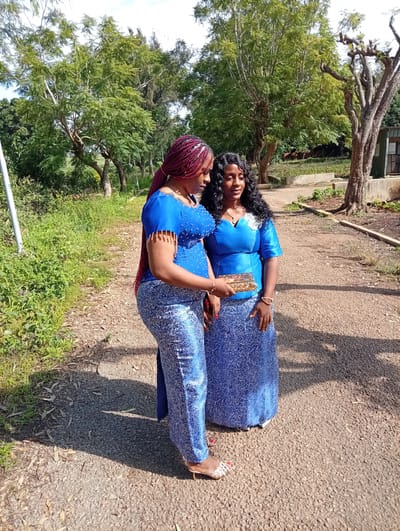 Two women are standing on a dirt path in a park-like setting. The woman on the left is wearing a blue beaded top and blue patterned pants, with red braided hair. She is holding a gold clutch. The woman on the right is wearing a blue dress with a white floral applique and has long, curly black hair. They are both looking at each other and smiling. The background consists of trees and green grass. The image appears to be taken in the city of Tambes Mile Four, Nigeria.