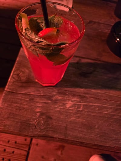A close-up, overhead shot shows a strawberry and mint infused beverage in a hexagonal glass, filled with ice and a bright red liquid. The glass is positioned on a rustic wooden table, with a dark background behind it. The lighting casts a warm, reddish hue over the entire scene. The location is identified as Oudenaarde, Belgium.