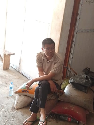 An adult male, appearing to be of East Asian descent, is seated on a stack of sacks within what appears to be an indoor or semi-enclosed, utilitarian space. He wears a light beige polo shirt, dark trousers, and green flip-flops, with clear-framed glasses. His arms are folded, and he is looking towards the right side of the frame. He sits on a light-colored sack, with an orange sack marked "VEER" partially visible beneath his legs. To his right, a pile of assorted sacks includes a grey sack and a red sack displaying the word "Brothers"; a plastic Coca-Cola bottle is visible partially obscured beneath the red sack. To his left, a plastic water bottle with a blue cap rests on the concrete floor. The background on the left features a brightly illuminated white wall or partition with white vertical framing, and a wooden bench is positioned further left. A brown structural pillar is visible on the right, with various objects, possibly metal poles, leaning against it. Faint handwritten text "Accepted" is visible on a surface in the upper right quadrant of the image. The specific location within Olomoda, Nigeria, cannot be determined from the visual evidence presented.
