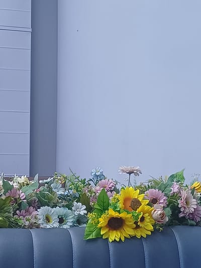 A close-up, vertically oriented, eye-level shot shows a floral arrangement atop a dark grey, tufted surface that appears to be a sofa. The flowers are artificial and include several sunflowers with dark centers and yellow petals, surrounded by a variety of other blooms in shades of pink, lavender, light blue, and cream, interspersed with green foliage. The background is a plain, light blue wall. To the left of the wall, a section of a light grey, ribbed wall or partition is visible. The scene is well-lit, suggesting an indoor setting. There are no people or animals present in the image. The specific location in Shahand, Uzbekistan, cannot be determined from the visual information provided.