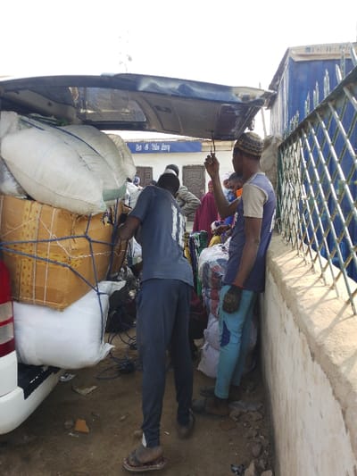 The image presents an outdoor scene in Unguwa Uku Kauyen Alu, Nigeria, focused on a white van with its rear hatch open and heavily loaded. The cargo consists of numerous large bundles, including white sacks and brown-wrapped packages, secured with blue rope and stacked above the vehicle's roofline.
Two adult males are prominent in the foreground. The male on the left, viewed from the rear, wears a dark blue t-shirt with a light-colored numeral on the back, dark blue trousers, and sandals. He is positioned at the rear of the van, interacting with the loaded items. The male on the right, viewed from the side, wears a dark blue and light blue t-shirt, light blue trousers, a patterned cap, and a dark glove on his right hand. He leans against a low concrete wall, holding an object connected to the van's open hatch.
In the mid-ground, behind the van, several indistinct individuals are visible. A building with a blue awning displaying "Eat & Take Away" in white text is present, featuring two brown doors below the awning. To the far right, a blue metal fence is situated atop the concrete wall, and a larger blue structure is visible beyond it. The ground is unpaved dirt with scattered debris. The scene is under bright daylight conditions, evident from distinct shadows.