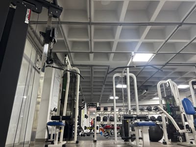 The image depicts the interior of a gym, likely in Boca del Río, Mexico, with exercise equipment positioned on a gray, waffle-patterned ceiling. Several weight machines are visible, along with rows of dumbbells and exercise balls in the background. Overhead wiring is present, and light fixtures illuminate the space. The BH FITNESS logo is prominently displayed on one of the machines.