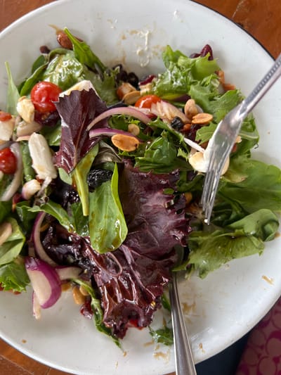 A close-up, overhead shot captures a salad in a white, circular bowl on a wooden surface. The salad is a mix of greens, including red leaf lettuce and other leafy vegetables. Visible ingredients include halved cherry tomatoes, slivers of red onion, white cheese cubes, dried cranberries, raisins, and nuts, possibly almonds or peanuts. A fork is actively engaged in the salad, lifting a portion to be eaten, with some dressing visible on the fork and in the bowl. The lighting is natural and appears to be daylight. The setting is a casual meal, likely in a restaurant or home in Chipilo de Francisco Javier Mina, Mexico, a town known for its Italian heritage and distinct cuisine.