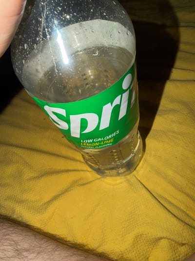 The image shows a partially full, plastic Sprite bottle lying on a crumpled yellow fabric surface, possibly a cushion or blanket. The bottle has condensation droplets on its exterior. Part of a person's arm with visible hair is visible on the right side of the frame. The location is not identifiable from the image.