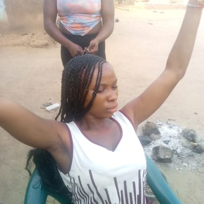 The scene is set outdoors in Ibreda, Nigeria. A dark-skinned female, seated in a dark green plastic chair, is having her hair braided by a second dark-skinned female standing directly behind her. The seated woman wears a white sleeveless V-neck top with brown vertical stripes on the lower portion and has numerous dark twist braids partially completed on her head. Her arms are raised and extended outwards, consistent with hair braiding. The standing woman wears a light-colored, tie-dye crop top and dark pants, with her hands engaged in the braiding process. The ground is composed of unpaved dirt or sand. To the right of the seated woman, an area of white ash and several dark rocks suggests the remains of a fire pit. Distant, blurred building structures are visible in the background.
